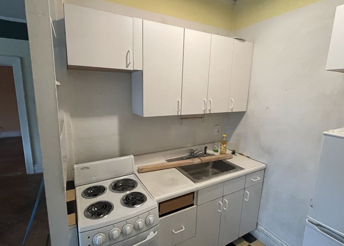Dated apartment kitchen with worn cabinets and checkerboard vinyl floor before renovation
