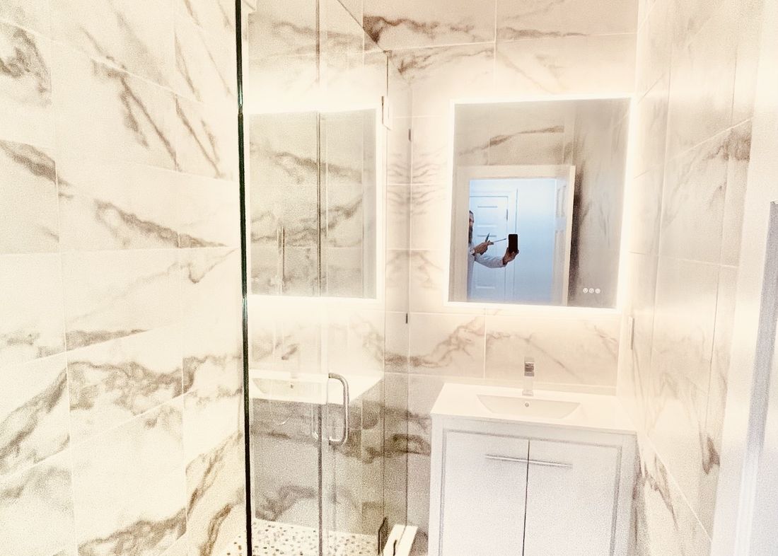 The same bathroom finished in marble tile with a frameless glass shower and backlit mirror